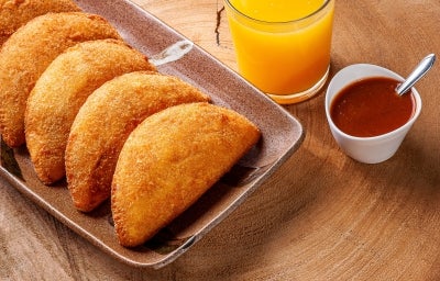 Fotografia de cinco risoles de presunto e queijo sobrepostos em um recipiente retangular marrom escuro sobre uma mesa de madeira. Ao lado dos salgados tem suco de laranja no copo de vidro e um molho vermelho em um recipiente branco pequeno. Fotografia de cinco risoles de presunto e queijo sobrepostos em um recipiente retangular marrom escuro sobre uma mesa de madeira. Ao lado dos salgados tem suco de laranja no copo de vidro e um molho vermelho em um recipiente branco pequeno.