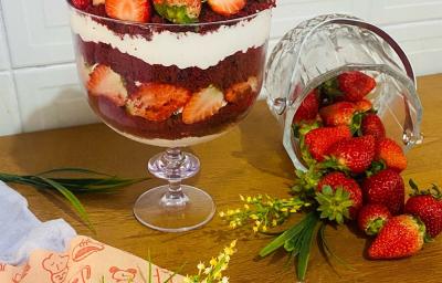 Foto em tons de vermelho da receita de red velvet na taça servida em uma graned porção em uma taça de vidro alta em cima de uma mesa de madeira com um pote de vidro com morangos ao lado Foto em tons de vermelho da receita de red velvet na taça servida em uma graned porção em uma taça de vidro alta em cima de uma mesa de madeira com um pote de vidro com morangos ao lado