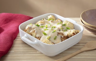 Fotografia em tons claros de uma recipiente quadrado de vidro e fundo sobre uma mesa de madeira clara. Dentro do recipiente tem a receita de couve flor e molho branco com um pouco de cebolinha picada por cima.