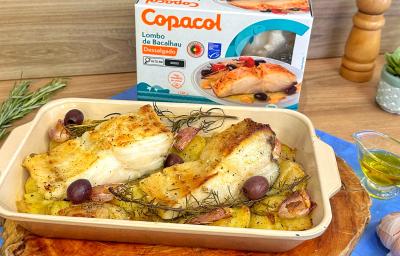Fotografia em tons de azul e castanho com uma travessa ao centro. Dentro da travessa existem dois lombos de bacalhau assados juntamente com batatas de tamanho pequeno. Ao fundo existe uma embalagem de bacalhau congelado da marca Copacol.