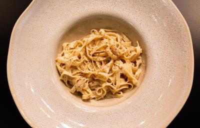 Fotografia em tons de cinza e preto com um prato cinza ao centro. Em cima do prato existe uma receita de cacio e pepe feita com macarrão tagliatelle e finalizado com pimenta preta moída Fotografia em tons de cinza e preto com um prato cinza ao centro. Em cima do prato existe uma receita de cacio e pepe feita com macarrão tagliatelle e finalizado com pimenta preta moída