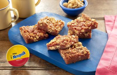 Fotografia em tons de marrom. em uma bancada de madeira de cor marrom. Ao centro, uma tábua de madeira azul contendo em cima, algumas fatias de brownies. Ao lado, um pano rosa e uma cápsula de Nesquik. Ao fundo, duas xícaras amarelas e um pote azul.