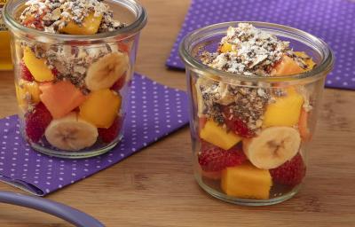 Fotografia de dois copos de vidro com frutas e granola sobre um paninho roxos com bolinhas brancas, sobre uma mesa de madeira.