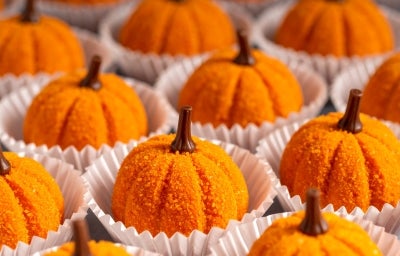 Fotografia em tons de laranja com vários brigadeiros em formato de abóbora ao centro. Os brigadeiros são feitos com abóbora cozida, passados no açúcar e finalizados com um cravo, para imitar a forma de uma abóbora.