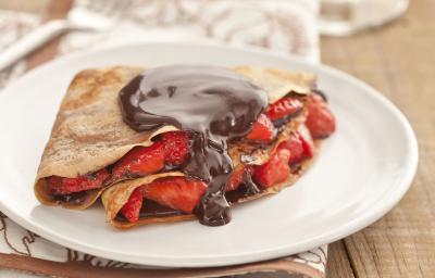 Fotografia em tons de marrom em uma bancada de madeira com um pano branco com listras marrom, um prato branco raso com o crepe recheado com morangos e chocolate. Fotografia em tons de marrom em uma bancada de madeira com um pano branco com listras marrom, um prato branco raso com o crepe recheado com morangos e chocolate.