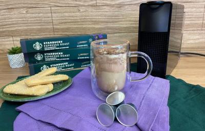 Fotografia em tons de verde e roxo em uma bancada de madeira, um paninho verde e roxo ao centro com uma xícara de vidro e o café tiramisu dentro. Ao fundo, uma máquina nespresso. Fotografia em tons de verde e roxo em uma bancada de madeira, um paninho verde e roxo ao centro com uma xícara de vidro e o café tiramisu dentro. Ao fundo, uma máquina nespresso.