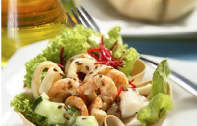 Fotografia em tons de verde em uma mesa com um vidro de azeite, um prato branco raso, um recipiente branco em formato de concha com a salada de alface, beterraba ralada, cenoura ralada e camarões dentro dele.