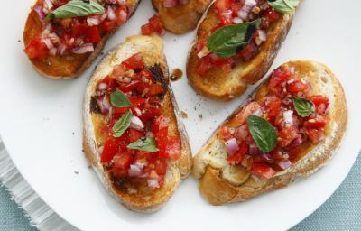 Fotografia vista de cima de cinco fatias de pão italiano torrado com uma bruschetta por cima. Os aperitivos estão sobre um prato de vidro branco, que está apoiado em um pano de mesa de cor azul.