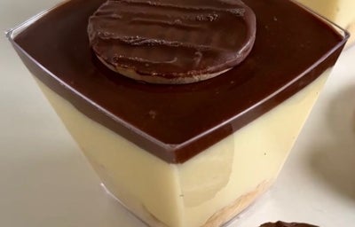 Fotografia em tons claros de dois potinhos de plástico quadrados com uma receita de torta holandesa sobre uma mesa branca.