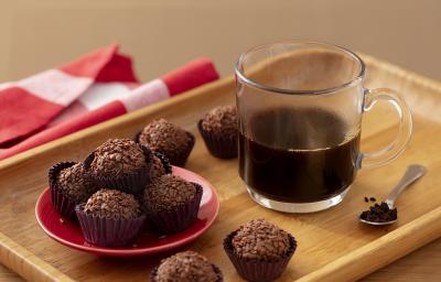 Fotografia em tons de marrom em uma bandeja de madeira de cor marrom. No centro, uma xícara de café preto e ao lado, alguns brigadeiros de Nescafé e uma colher contendo pó de Nescafé. Ao fundo, um pano vermelho. Fotografia em tons de marrom em uma bandeja de madeira de cor marrom. No centro, uma xícara de café preto e ao lado, alguns brigadeiros de Nescafé e uma colher contendo pó de Nescafé. Ao fundo, um pano vermelho.