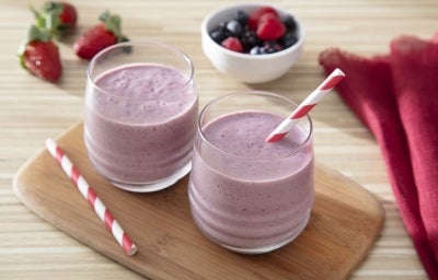 Fotografia de dois copos de vidro pequenos com o smoothie, em um deles têm um canudo vermelho e branco, o outro canudo está ao lado dos copos sobre uma mini tábua de madeira decorativa. A tábua está sobre uma mesa de madeira clara. Fotografia de dois copos de vidro pequenos com o smoothie, em um deles têm um canudo vermelho e branco, o outro canudo está ao lado dos copos sobre uma mini tábua de madeira decorativa. A tábua está sobre uma mesa de madeira clara.