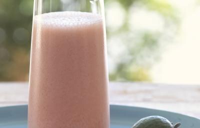 Fotografia de uma vitamina de goiaba com leite e neston com uma fatia fina de uma rodela de fruta na borda do copo de vidro. A bebida está apoiada em um prato azul, sobre uma mesa de madeira.