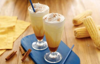 Fotografia em tons de amarelo, em uma bancada de madeira de cor marrom. Ao centro, uma tábua azul contendo 2 copos de milkshake. Ao lado, algumas canelas em pau espalhadas e ao fundo, um pano amarelo e algumas espigas de milho espalhadas. Fotografia em tons de amarelo, em uma bancada de madeira de cor marrom. Ao centro, uma tábua azul contendo 2 copos de milkshake. Ao lado, algumas canelas em pau espalhadas e ao fundo, um pano amarelo e algumas espigas de milho espalhadas.