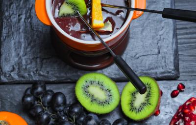 Fotografia de um Fondue de chocolate meio amargo dentro de um recipiente de vidro na cor laranja, com frutas sobre o creme. Ao lado, algumas frutas sobre uma mesa preta, como uvas roxas e um kiwi cortado na metade. Fotografia de um Fondue de chocolate meio amargo dentro de um recipiente de vidro na cor laranja, com frutas sobre o creme. Ao lado, algumas frutas sobre uma mesa preta, como uvas roxas e um kiwi cortado na metade.