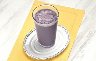 Fotografia em tons de roxo e amarelo em uma bancada de madeira clara, um pano amarelo, um prato branco redondo com um copo de vidro alto e dentro dele a bebida de uva com abacaxi. Fotografia em tons de roxo e amarelo em uma bancada de madeira clara, um pano amarelo, um prato branco redondo com um copo de vidro alto e dentro dele a bebida de uva com abacaxi.
