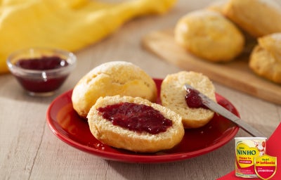 Pãezinhos abertos com geleia vermelha sobre prato vermelho, faca ao lado e mais pães ao fundo, em mesa clara.