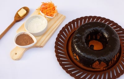 Um bolo de cenoura inteiro, com cobertura de chocolate brilhante cobrindo toda a superfície. Ao lado, há uma tábua com ingredientes usados na receita: manteiga, farinha, cacau em pó e cenoura ralada.