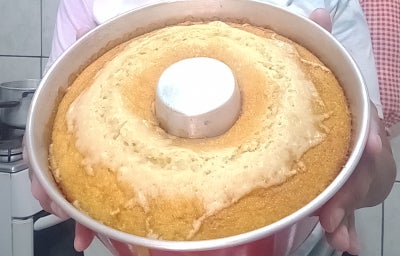 Bolo caseiro dourado em forma com furo central, recém-assado, sendo segurado em uma cozinha.