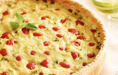 Fotografia em tons de amarelo e vermelho em uma bancada de madeira com uma toalha bege. Ao centro, o quiche de tomates com manjericão. Fotografia em tons de amarelo e vermelho em uma bancada de madeira com uma toalha bege. Ao centro, o quiche de tomates com manjericão.