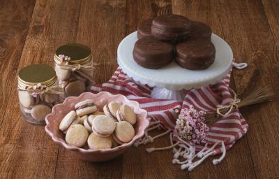 Foto da receita de alfajor com doce de leite, em diversos formatos redondos de tamanhos menor e maior, servidos sobre uma bancada de madeira decora com um tecido vermelho e branco listrado