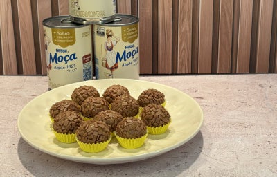 Prato com docinhos cobertos com granulado de chocolate, em forminhas amarelas, e latas de Leite Moça ao fundo.