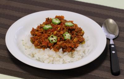 Fotografia em tons de marrom com um prato branco fundo ao centro. Dentro do prato existe uma porção de arroz coberta com carne moída e pedaços de quiabo. Ao lado existe uma colher. Fotografia em tons de marrom com um prato branco fundo ao centro. Dentro do prato existe uma porção de arroz coberta com carne moída e pedaços de quiabo. Ao lado existe uma colher.