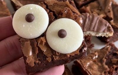Fotografia de uma mão masculina segurando o brownie de morcego. Ao fundo tem mais pedaços do brownie sobre um prato raso branco. Fotografia de uma mão masculina segurando o brownie de morcego. Ao fundo tem mais pedaços do brownie sobre um prato raso branco.
