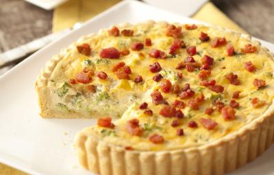 Fotografia em tons de marrom, branco e amarelo de uma bancada de madeira com um paninho amarelo, sobre ele um prato quadrado com um quiche e uma espátula. Ao fundo um prato quadrado branco com uma fatia de quiche.