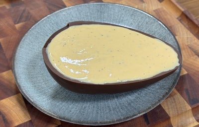 imagem mostra um ovo de páscoa de chocolate escuro, ao meio, recheado de um creme amarelado e polpa de maracujá por cima