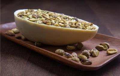 Fotografia de uma metade de ovo de páscoa sobre um apoio retangular de madeira, com alguns pistaches espalhados ao lado. O ovo de páscoa tem a casca de chocoalte branco, o recheio de brigadeiro de pistache e pistaches por cima.