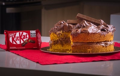 Fotografia em tons escuros de um bolo de cenoura com ganache de kitkat no recheio e na cobertura. As laterais do bolo estão sem a ganache, o bolo está apoiado em um pano vermelho sobre uma mesa cinza. Ao lado tem uma embalagem de kitkat. Fotografia em tons escuros de um bolo de cenoura com ganache de kitkat no recheio e na cobertura. As laterais do bolo estão sem a ganache, o bolo está apoiado em um pano vermelho sobre uma mesa cinza. Ao lado tem uma embalagem de kitkat.