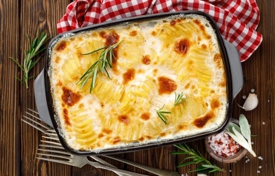 Em cima de uma mesa, refratário com batatas gratinadas com molho branco, decorado com alecrim.