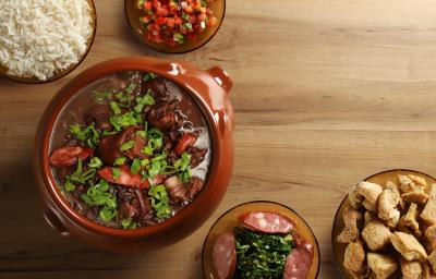 Fotografia vista de cima de uma feijoada completa, com cebolinha por cima, em um recipiente grande clássico de vidro marrom. Ao redor tem acompanhamentos de arroz, torresmo, vingrete e couve, todos em um recipiente de vidro, sobre uma mesa de madeira Fotografia vista de cima de uma feijoada completa, com cebolinha por cima, em um recipiente grande clássico de vidro marrom. Ao redor tem acompanhamentos de arroz, torresmo, vingrete e couve, todos em um recipiente de vidro, sobre uma mesa de madeira