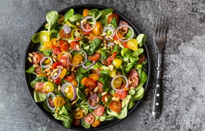 Fotografia vista de cima de uma salada de milho e pequi com folhas, tomates cereja, cebola roxa e grãos em um prato raso preto. Ao lado da salada, um garfo com cabo de cor preta, sobre uma mesa cinza. Fotografia vista de cima de uma salada de milho e pequi com folhas, tomates cereja, cebola roxa e grãos em um prato raso preto. Ao lado da salada, um garfo com cabo de cor preta, sobre uma mesa cinza.