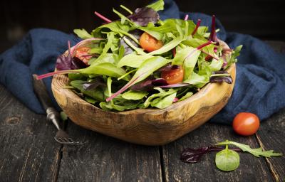 Fotografia em tons escuros de uma salada verde com salsão e tomates cereja dentro de um recipiente fundo de madeira, que está apoiado em um pano azul escuro, juntamente com um garfo de cabo preto, sobre uma mesa de madeira escura. Fotografia em tons escuros de uma salada verde com salsão e tomates cereja dentro de um recipiente fundo de madeira, que está apoiado em um pano azul escuro, juntamente com um garfo de cabo preto, sobre uma mesa de madeira escura.