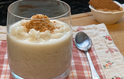 Imagem de um copo transparente ao centro com uma porção de arroz doce branco cremoso com uma farofa de biscoio triturado dourada por cima. o Copo está apoiado em cima de um pano xadrez vermelho e branco. Ao fundo, um biscoito Tostines.