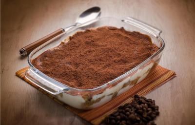 Fotografia de uma torta de bolacha com nata polvilhada com cacau em pó. A torta está em um recipiente refratário retangular de vidro que está sobre um pano marrom sobre uma mesa de madeira. Fotografia de uma torta de bolacha com nata polvilhada com cacau em pó. A torta está em um recipiente refratário retangular de vidro que está sobre um pano marrom sobre uma mesa de madeira.