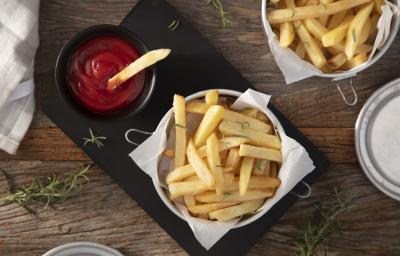 Fotografia em tons de marrom em uma bancada de madeira escura, uma tábua de ferro preta com um pote com batatas fritas dentro e um potinho ao lado com ketchup. Ao lado, um paninho e mais batata frita. Fotografia em tons de marrom em uma bancada de madeira escura, uma tábua de ferro preta com um pote com batatas fritas dentro e um potinho ao lado com ketchup. Ao lado, um paninho e mais batata frita.