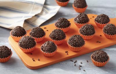 Imagem aproximada da receita de Brigadeiro Zero Lactose. Há diversos brigadeiros enrolados em forminhas em tom de laranja, todos sobre uma tábua de madeira em uma bancada decorada com um tecido em tom azul e bege Imagem aproximada da receita de Brigadeiro Zero Lactose. Há diversos brigadeiros enrolados em forminhas em tom de laranja, todos sobre uma tábua de madeira em uma bancada decorada com um tecido em tom azul e bege