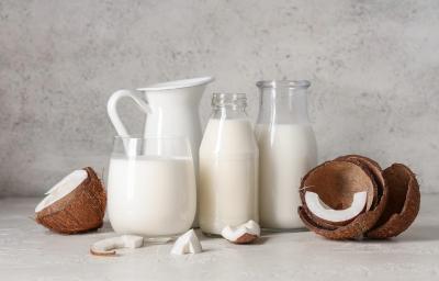 Fotografia de duas garrafas decorativas de vidro, uma jarra branca e um copo grande e largo. Todos esses recipientes têm bebida vegetal de coco. Ao lado tem cascas de coco pela metade, alguns pedaços e um pouco da polpa sobre uma mesa cinza clara. Fotografia de duas garrafas decorativas de vidro, uma jarra branca e um copo grande e largo. Todos esses recipientes têm bebida vegetal de coco. Ao lado tem cascas de coco pela metade, alguns pedaços e um pouco da polpa sobre uma mesa cinza clara.