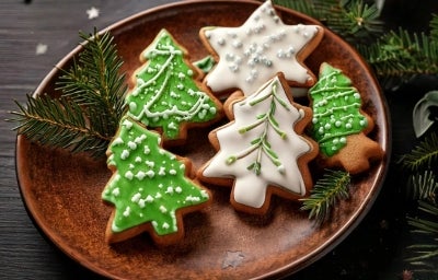 Em cima de uma mesa, prato redondo com biscoitos em forma de árvore de Natal e estrela, decorado com glacê branco e verde, e confeitos redondos brancos. Em cima de uma mesa, prato redondo com biscoitos em forma de árvore de Natal e estrela, decorado com glacê branco e verde, e confeitos redondos brancos.