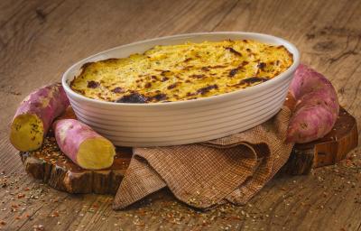 Fotografia em tons de marrom em uma bancada de madeira marrom. No centro, há um pano marrom com uma travessa branca em cima contendo o Escondidinho de Batata Doce com algumas batatas doces ao redor. Fotografia em tons de marrom em uma bancada de madeira marrom. No centro, há um pano marrom com uma travessa branca em cima contendo o Escondidinho de Batata Doce com algumas batatas doces ao redor.