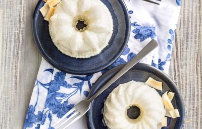 Fotografia em tons de azul em uma bancada de madeira clara, um pano branco com folhas azuis e dois pratinhos azuis escuros com o mini pudim de tapioca feito com iogurte, abacaxi e coco.