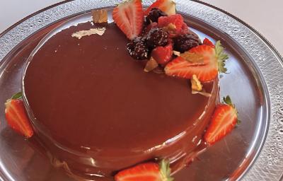 Fotografia em tons de vermelho e marrom com um prato prateado ao centro. Em cima do prato existe um bolo de chocolate coberto com calda de chocolate e frutas vermelhas
