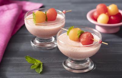 Fotografia em tons de rosa em uma bancada de madeira escura, um paninho rosa, duas taças de vidro com o creme de iogurte de morando com espetinhos de fruta em cima dele.