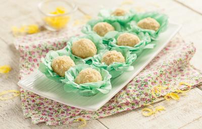 Fotografia em tons de verde em uma bancada de madeira com um recipiente branco retangular com os brigadeiros de torta de limão em forminhas verdes colocados em cima do recipiente. Ao lado, raspas de limão-siciliano espalhadas pela mesa. Fotografia em tons de verde em uma bancada de madeira com um recipiente branco retangular com os brigadeiros de torta de limão em forminhas verdes colocados em cima do recipiente. Ao lado, raspas de limão-siciliano espalhadas pela mesa.