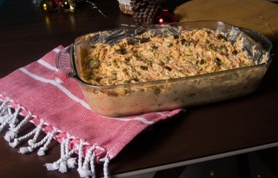 Fotografia de um prato de salpicão cremoso em uma travessa de vidro, com cenouras raladas, passas, azeitonas e um toque festivo, decorado com um pano vermelho e branco, sugerindo um ambiente acolhedor e de celebração.
