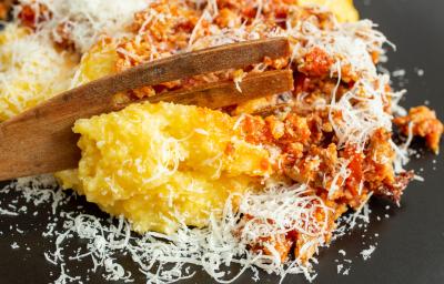 Fotografia de um prato raso preto com uma polenta gratinada recehada com carne moída e queijo ralado por cima. Apoiado na receita, tem um talher de madeira grande.