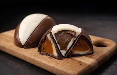 Fotografia em tons de marrom em uma bancada escura e uma tábua de madeira ao centro. Em cima, ovos de Páscoa cortados em fatias com recheios dentro.