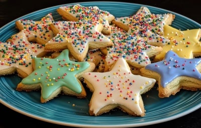 Em cima de uma mesa, prato redondo com biscoitos em formato de estrelas de Natal, decorados com glacê e confeitos de bolinhas coloridas. Em cima de uma mesa, prato redondo com biscoitos em formato de estrelas de Natal, decorados com glacê e confeitos de bolinhas coloridas.